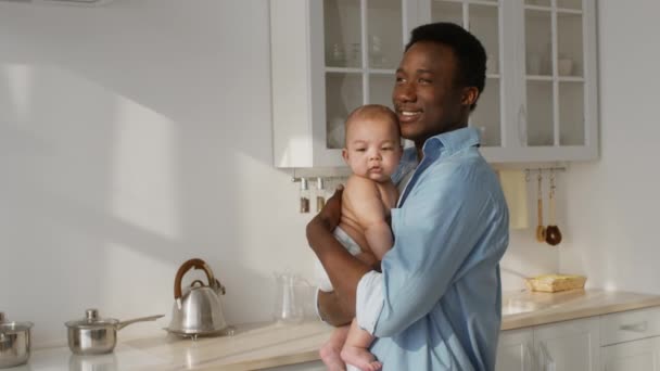 Positive african american dad carrying his newborn baby and looking aside, enjoying early morning at kitchen — Stock videók