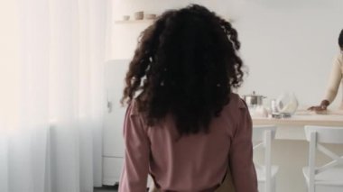 Follow shot of african american girl going to mother and helping her, adding flour to dough at kitchen, slow motion