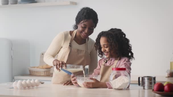 Happy african american mother and daughter preparing dough, woman adding milk, girl whisking it in bowl at kitchen — стокове відео