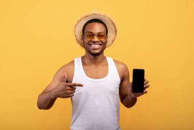 Yazlık kıyafetli siyahi adam, sarı arka planda boş ekranlı cep telefonuna bakıyor. Model için yer var.