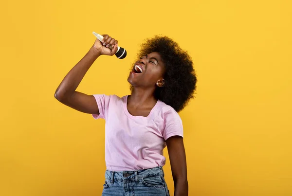 Alegre dama afroamericana cantando canciones en micrófono, divirtiéndose y disfrutando del momento, fondo amarillo —  Fotos de Stock