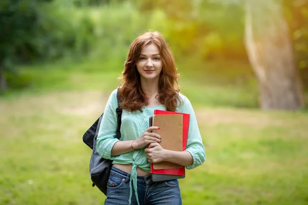 Привлекательная рыжая студентка с рюкзаком и рабочими тетрадями, позирующая на территории кампуса — стоковое фото