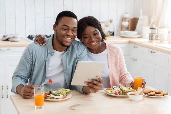 Loisirs domestiques. Couple africain mangeant le petit déjeuner et utilisant la tablette numérique dans la cuisine — Photo