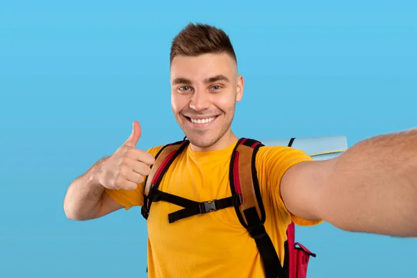 Feliz viajante masculino com mochila mostrando polegar para cima gesto ao fazer selfie sobre fundo estúdio azul — Fotografia de Stock