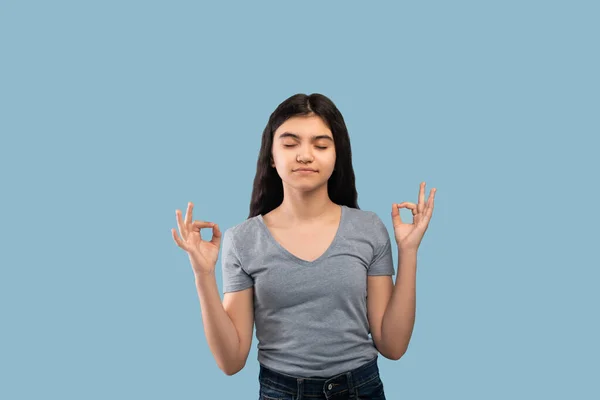 Peaceful Indian teen girl meditating with closed eyes over blue studio background — Stock Photo, Image