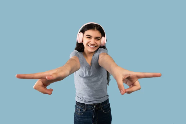 India adolescente chica en casual desgaste usando auriculares inalámbricos, señalando los dedos en diferentes lados en fondo azul —  Fotos de Stock