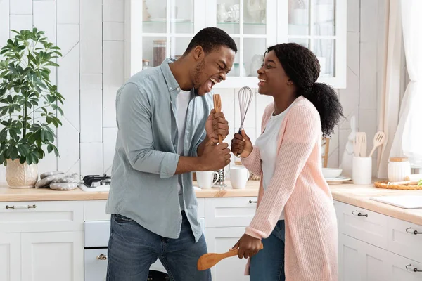 Concert domestique. drôles noir conjoints tromper dans cuisine, chanter avec des ustensiles — Photo