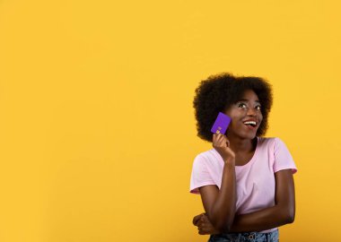 Alışveriş zamanı. Hayalperest Afro-Amerikalı kadın kredi kartı tutuyor ve başka tarafa bakıyor, sarı arka plan ve fotokopi alanı