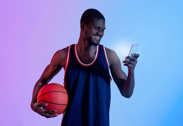 Penggemar bola basket hitam positif menonton kejuaraan online, bersorak untuk tim favoritnya di smartphone dalam cahaya neon — Stok Foto