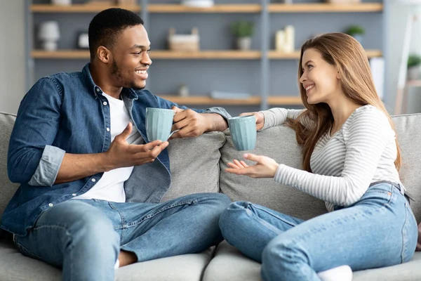 Wesoła mieszana para relaksująca się na kanapie w domu z kawą i rozmową — Zdjęcie stockowe