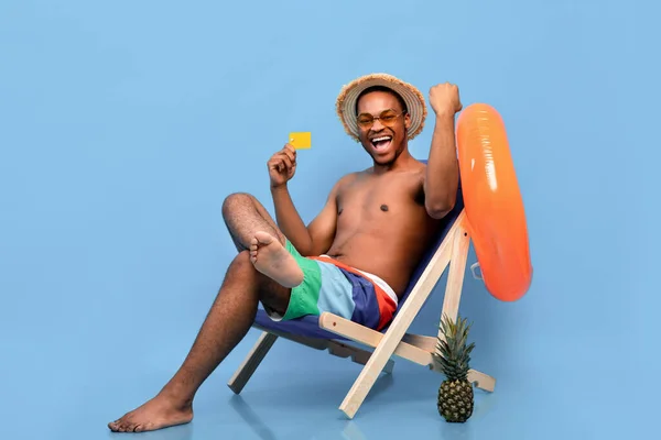 Emocionado hombre negro en traje de baño con tarjeta de crédito, sentado en el sillón, haciendo un gesto SI sobre fondo azul —  Fotos de Stock