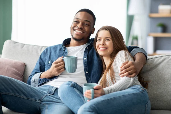 Aile içi hafta sonu. Mutlu çok kültürlü çiftler televizyon izleyip evde kahve içiyorlar. — Stok fotoğraf