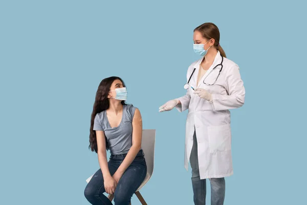Doctora joven hablando con paciente adolescente después de aplicarle la vacuna contra el coronavirus sobre fondo azul del estudio — Foto de Stock