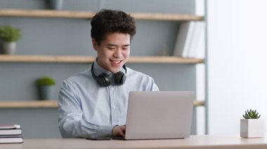 Love my job. Cheerful asian man with headphones on neck typing on laptop, working remotely at IT department