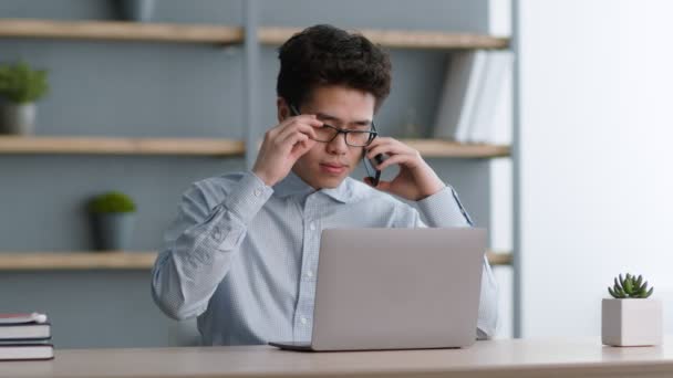Consulta remota. Jovem gerente asiático trabalhando no laptop e chamando fornecedores, discutindo negócios via celular — Vídeo de Stock