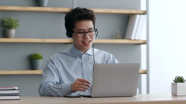 Glada asiatiska killen bär headset prata med kollegor, diskutera affärer och marknadsföring strategi, sitter hemma — Stockvideo