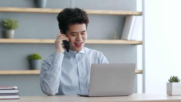 Bilgisayarda çalışan ve akıllı telefondan müşteriyle konuşan pozitif Asyalı yönetici, bilgisayardaki bilgileri kontrol ediyor — Stok video