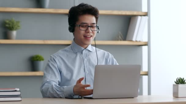 Pozytywne młody azjatycki kierownik biura noszenie słuchawki rozmowy z współpracownikami, przekazywanie wiadomości do rozmówców za pośrednictwem laptopa — Wideo stockowe