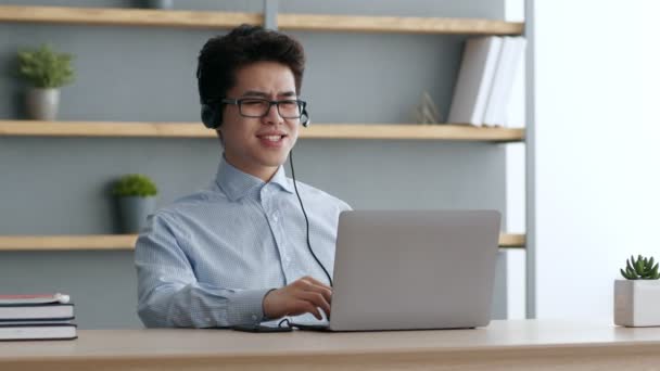 Iloinen aasialainen johtaja yllään kuulokkeet nauraa työpaikalla, video chattailuun kollegoiden kanssa etänä, työskentelevät toimistossa — kuvapankkivideo