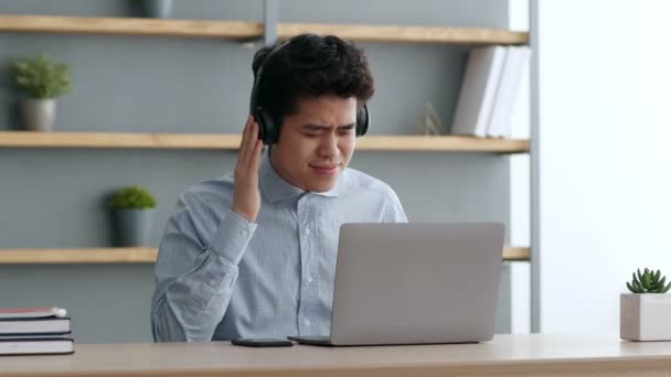 Asiatisk kontorsarbetare med hörlurar som njuter av musik på nätet, dansar på arbetsplatsen medan han arbetar med bärbar dator, slow motion — Stockvideo