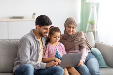 Genç Müslüman ebeveynler ve kız birlikte dizüstü bilgisayar kullanıyorlar, internette geziniyorlar veya biriyle görüntülü sohbet ediyorlar, kanepede oturuyorlar.