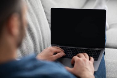 Kanepeye yaslanmış bir adam, boş ekranlı dizüstü bilgisayar kullanıyor.