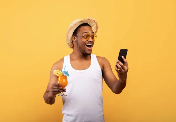 Fiatal fekete srác koktél nézi az okostelefon izgalomban, nyertes lottó vagy kaszinó, sárga háttér — Stock Fotó