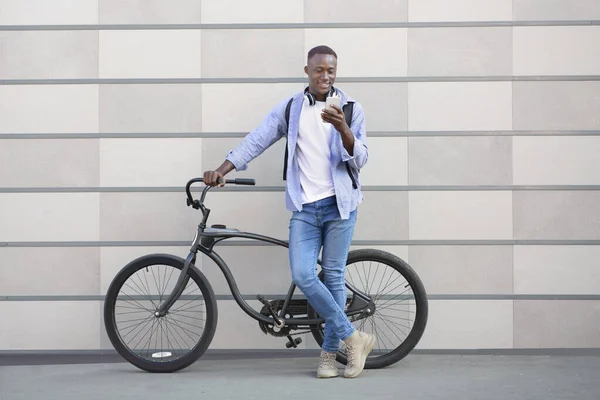 Homem afro-americano alegre com bicicleta de pé perto da parede de tijolo e escolhendo playlist de música no smartphone — Fotografia de Stock