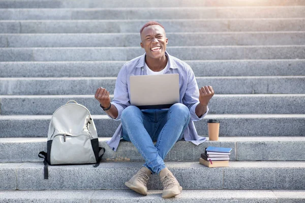 Remote education concept. Excited young black man with laptop computer gesturing YES on stairs outdoors