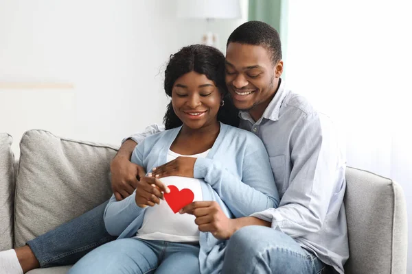 Fröhlich erwartetes Paar sitzt auf Couch und hält rotes Herz — Stockfoto