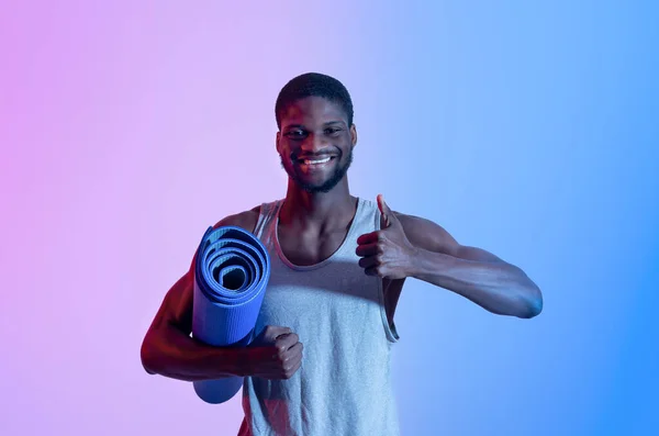 Pria kulit hitam muda yang bahagia memegang tikar yoga, menunjukkan gerakan jempol dalam pencahayaan neon — Stok Foto
