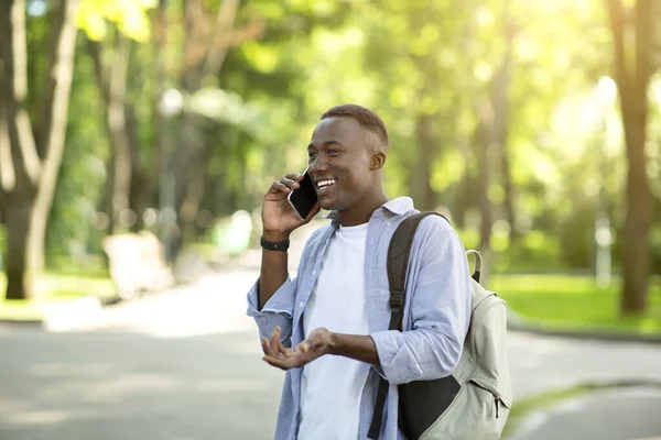 Cara preto feliz com mochila falando no telefone celular no parque da cidade, espaço livre — Fotografia de Stock