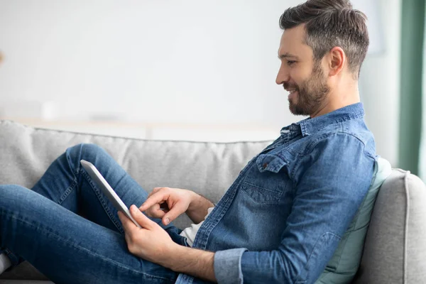 Homem feliz reclinado no sofá em casa, usando tablet digital — Fotografia de Stock