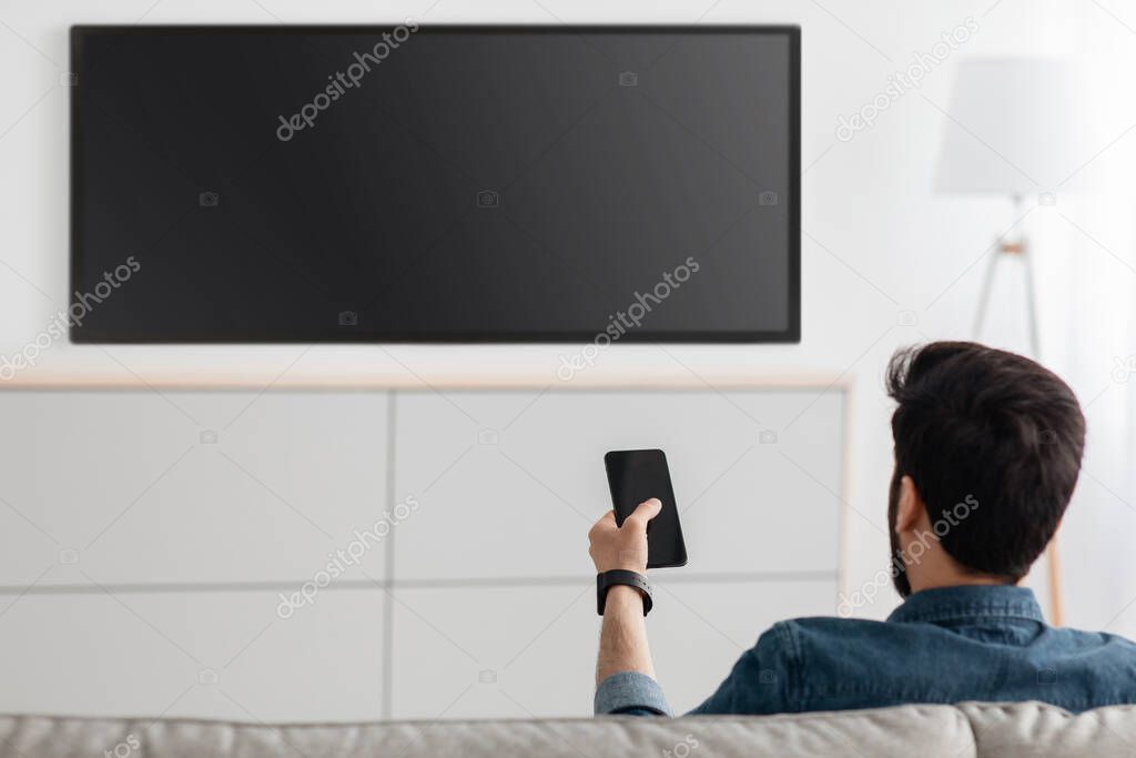 Young man watching television and using smart TV remote control application on smartphone, back view