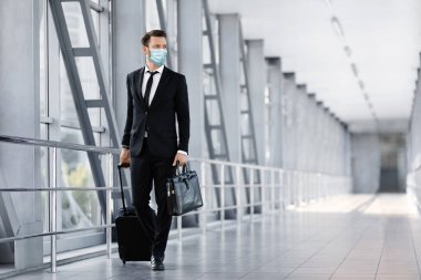 Yüzü maskeli bir iş adamı havaalanı terminalinde yürüyor.