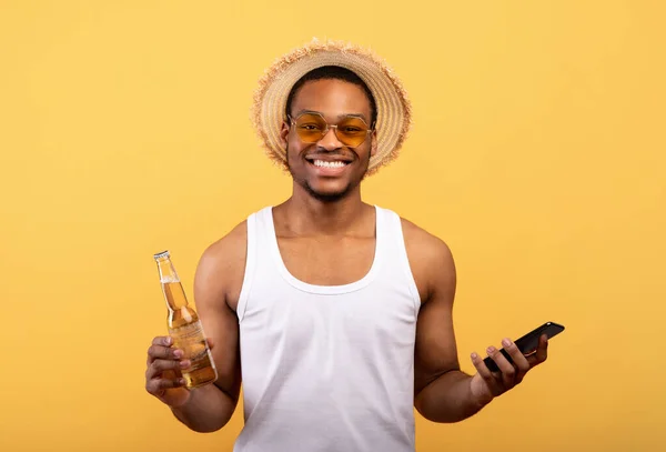 Vidám afro-amerikai srác gazdaság üveg sör és okostelefon sárga stúdió háttér — Stock Fotó