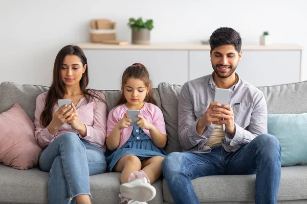 Concepto de adicción al gadget. Familia árabe joven de tres personas que sostiene y utiliza teléfonos inteligentes mientras está sentado en el sofá en la sala de estar — Foto de Stock