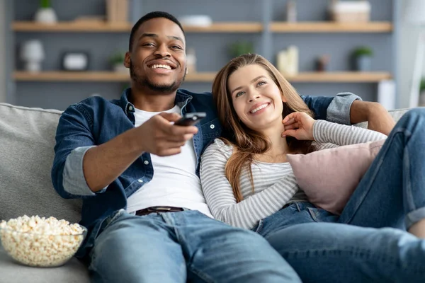 Passatempo doméstico. Casal de raça mista feliz com pipoca assistindo TV em casa — Fotografia de Stock