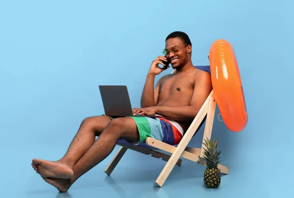 Trabalho on-line em férias de verão. Preto cara com laptop falando no smartphone enquanto sentado na cadeira lounge — Fotografia de Stock