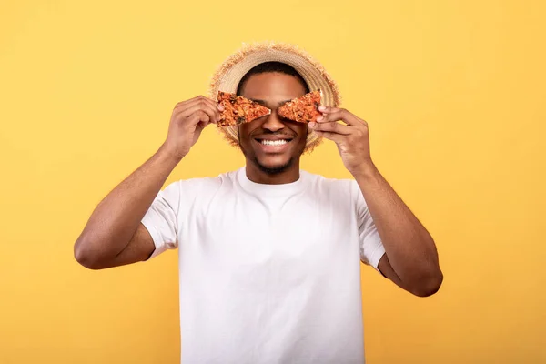 Retrato de cara preto feliz cobrindo seus olhos com fatias de pizza gostosa no fundo do estúdio amarelo — Fotografia de Stock