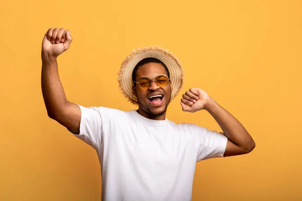 Bonito homem negro de óculos de sol, t-shirt e chapéu de palha levantando as mãos, dançando no fundo do estúdio amarelo — Fotografia de Stock