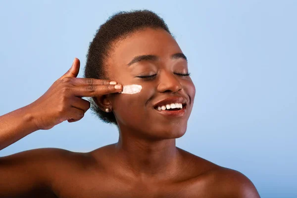 Mulher negra feliz rosto hidratante, aplicando creme na bochecha, posando com os olhos fechados no fundo azul — Fotografia de Stock