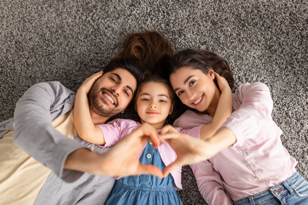 Feliz família amorosa. Pais árabes e filha liitle deitado no tapete de chão e de mãos dadas em forma de coração sinal — Fotografia de Stock