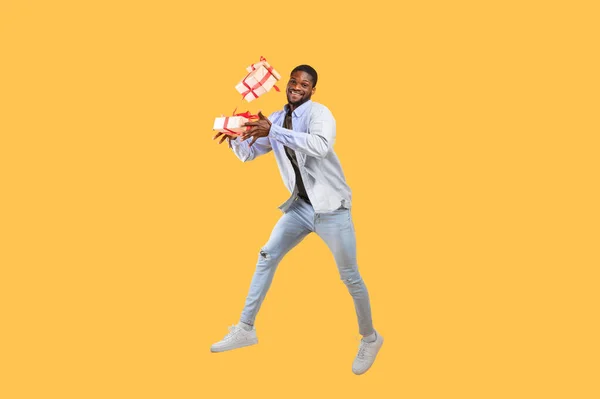 Días festivos y tiempo de celebración. Alegre afroamericano chico sosteniendo cajas de regalo y saltando sobre fondo amarillo — Foto de Stock