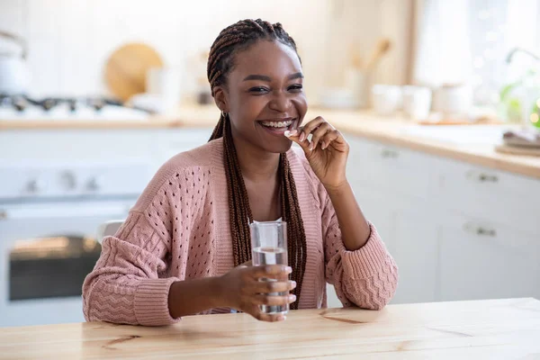 Alegre joven afroamericana dama tomando suplemento de belleza píldora en la cocina —  Fotos de Stock