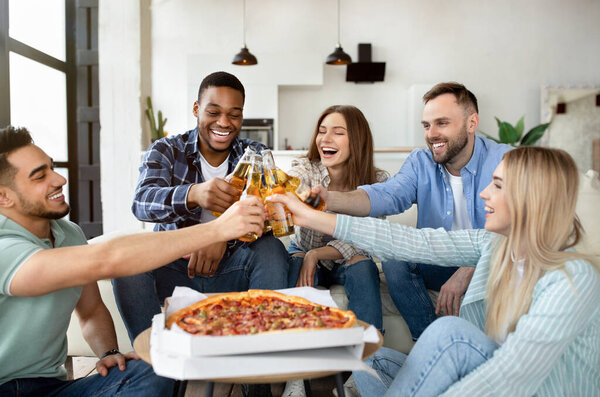 Carefree group of happy friends enjoying party, eating pizza, toasting with beer bottles, celebrating something at home