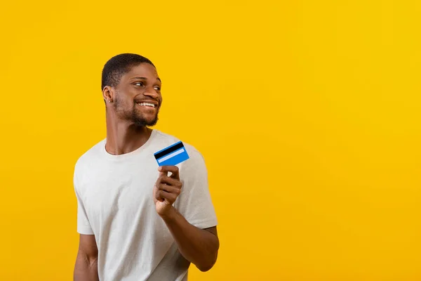 Emocionado hombre afroamericano con tarjeta de crédito y mirando el espacio vacío en el fondo del estudio amarillo —  Fotos de Stock
