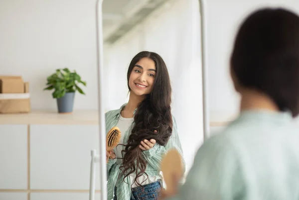 Veselý indický žena kartáčování její dlouhé krásné vlasy s dřevěným štětcem před zrcadlem doma — Stock fotografie