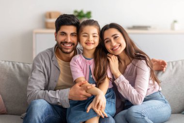 Aile fotoğrafı. Heyecanlı Arap ebeveynler ve sevimli küçük kız birlikte poz veriyor, kameraya sarılıyor ve gülümsüyor.