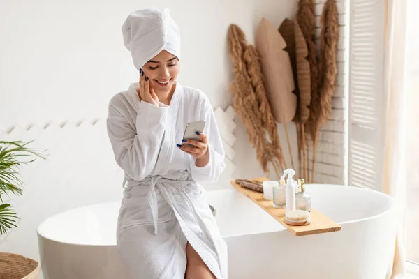 Mujer usando Smartphone en baño usando albornoz sentado en la bañera —  Fotos de Stock